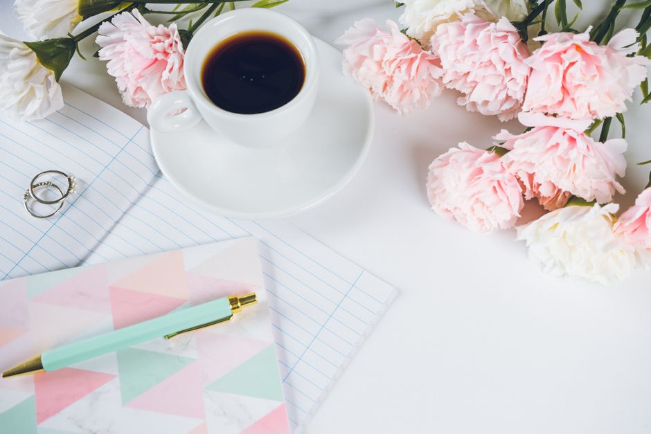 A stylish flat lay with coffee, peonies, notebook, and pen on a white desk.